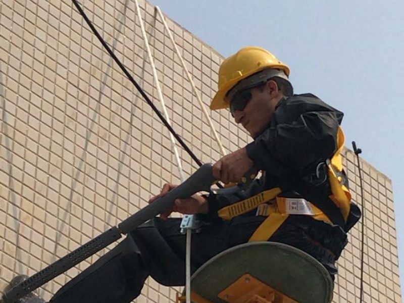 Alpinista industrial da HG Engenharia com capacete, óculos e cinto de segurança em cadeira suspensa na fachada de um edifício em Florianópolis - SC